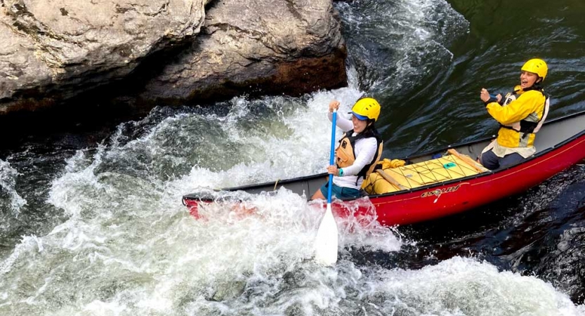 Two people wearing safety gear paddle a canoe through whitewater.